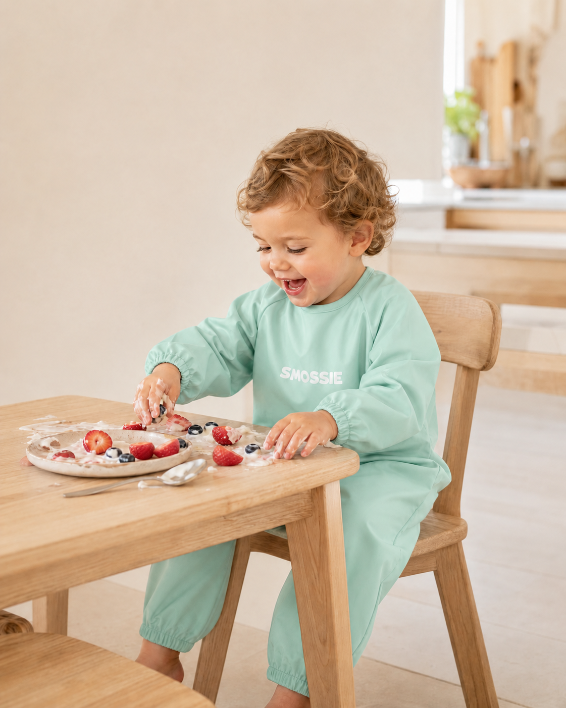 A toddler eating pancakes and yoghurt in a pink Smossie suit